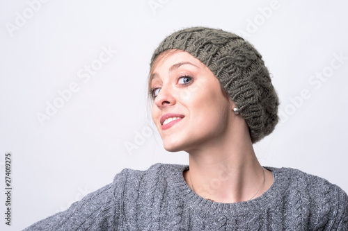 Portrait of confident charming young hipster woman with knitted cap posing and smiling on light gray background with copyspace. 