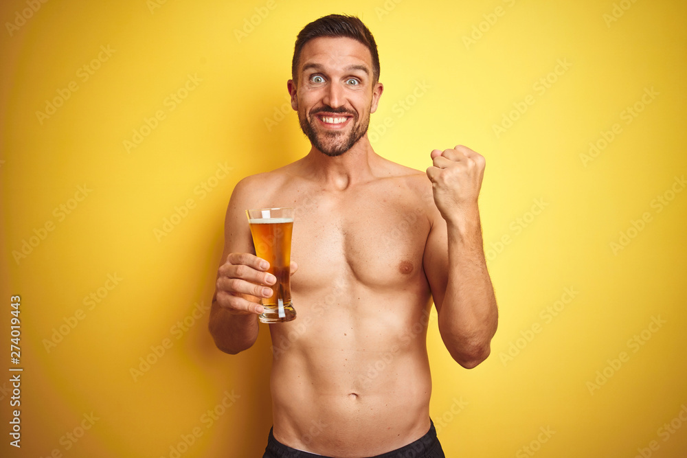 Fototapeta premium Young handsome shirtless man drinking a pint of beer over isolated yellow background screaming proud and celebrating victory and success very excited, cheering emotion