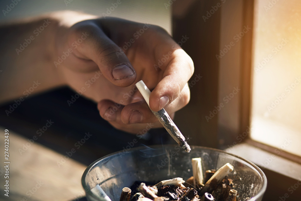 The man's hand holds a Smoking cigarette in his fingers and shakes the ashes from it into a dirty glass ashtray, standing on the windowsill and illuminated by sunlight.