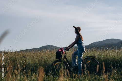 Silhouette girl with long curly hair in hat driving a bike on nature. She holds a bicycle with a basket of flowers and expressing happy emotions during active rest in weekend