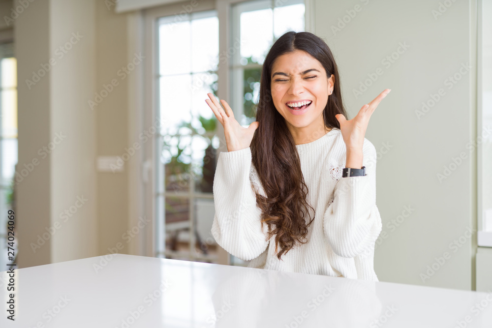 Young beautiful woman at home on white table celebrating mad and crazy for success with arms raised and closed eyes screaming excited. Winner concept Young beautiful woman at home on white table celebrating mad and crazy for success with arms raised and closed eyes screaming excited. Winner concept