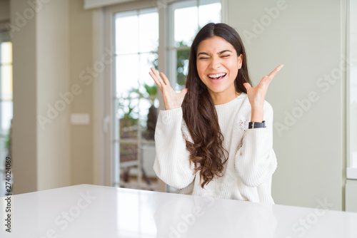 Young beautiful woman at home on white table celebrating mad and crazy for success with arms raised and closed eyes screaming excited. Winner concept