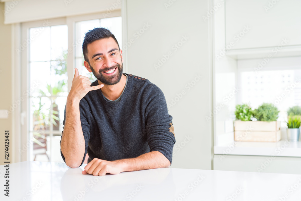 Handsome hispanic man wearing casual sweater at home smiling doing phone gesture with hand and fingers like talking on the telephone. Communicating concepts.