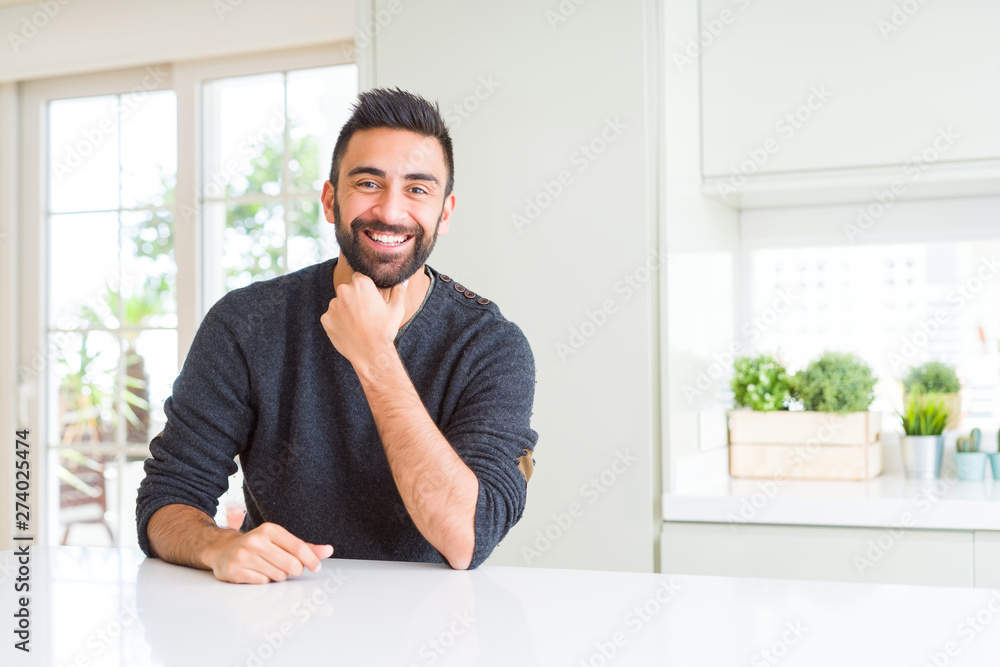 Handsome hispanic man wearing casual sweater at home looking confident at the camera with smile with crossed arms and hand raised on chin. Thinking positive.