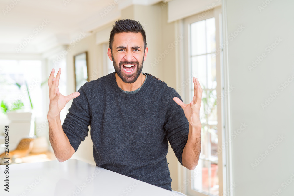 Handsome hispanic man wearing casual sweater at home crazy and mad shouting and yelling with aggressive expression and arms raised. Frustration concept. Handsome hispanic man wearing casual sweater at home crazy and mad shouting and yelling with aggressive expression and arms raised. Frustration concept.