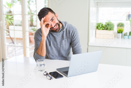 Handsome hispanic man working using computer laptop with happy face smiling doing ok sign with hand on eye looking through fingers