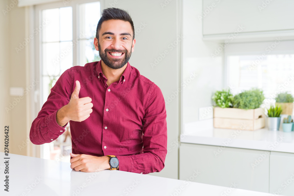 Handsome hispanic business man doing happy thumbs up gesture with hand. Approving expression looking at the camera showing success.