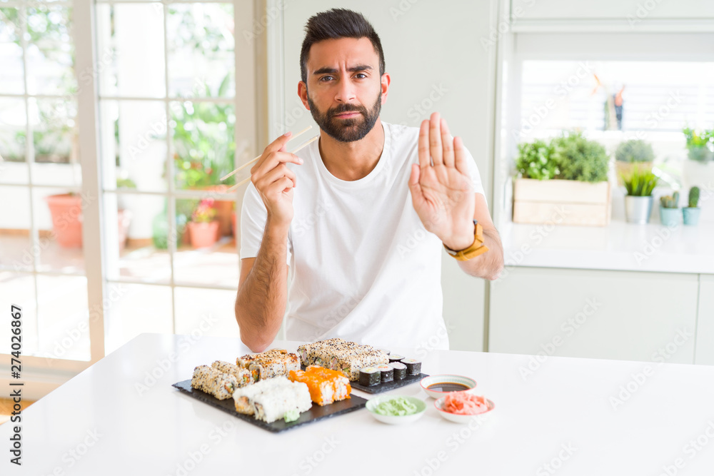 Handsome hispanic man eating asian sushi using chopsticks with open hand doing stop sign with serious and confident expression, defense gesture Handsome hispanic man eating asian sushi using chopsticks with open hand doing stop sign with serious and confident expression, defense gesture