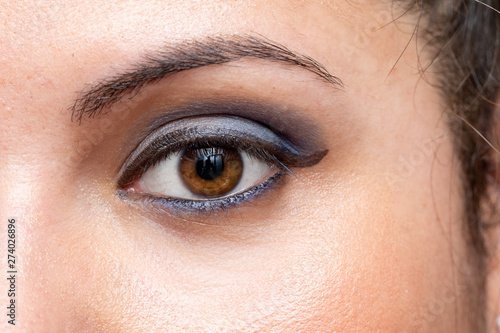 An extreme closeup view on the eye of a young Caucasian lady with brown iris looking to camera. Flawless and firm skin is seen beneath the eye as she wears smoky blue eyeshadow