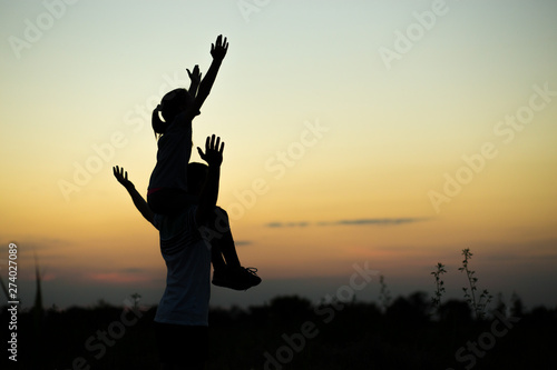 Silhouettes of father and daughter on his shoulders with hands up having fun, against sunset sky. Parenthood, family activities, beach holiday and vacation, support and love themes