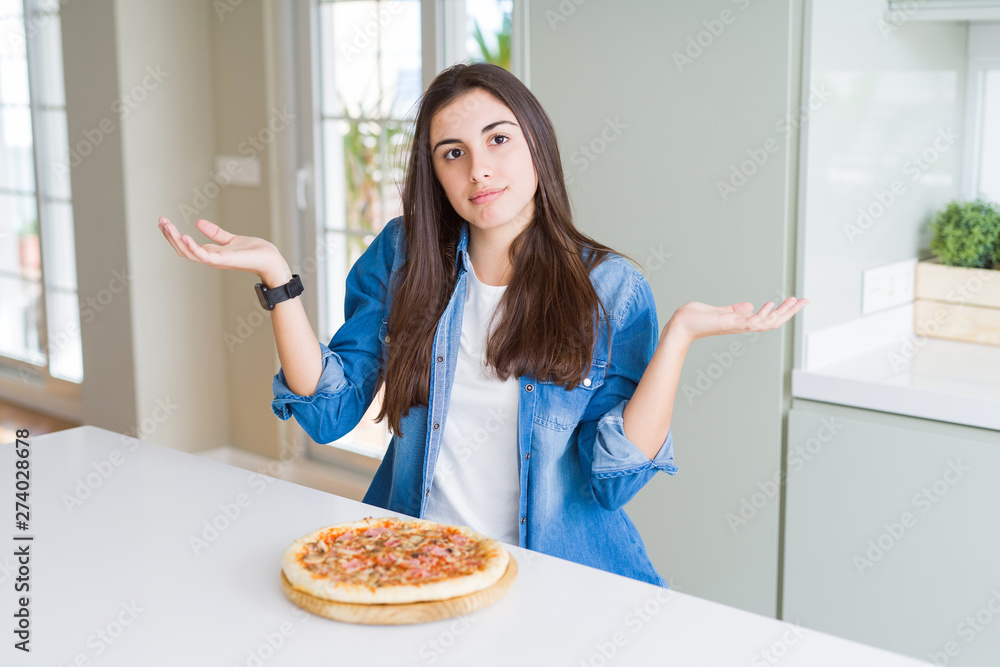 Beautiful young woman eating homemade tasty pizza at the kitchen clueless and confused expression with arms and hands raised. Doubt concept.