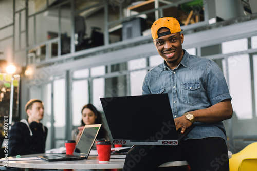 Fotografija Dark skinned African american coworker in jeans shirt and yellow cap and using laptop in modern office