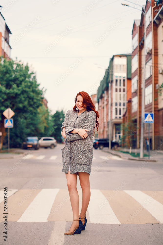 Red-haired Model in a coat posing against the backdrop of the sunset on the street.