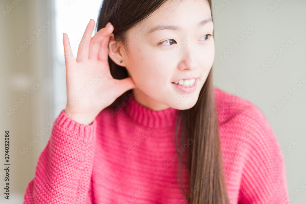 Beautiful Asian woman wearing pink sweater smiling with hand over ear listening an hearing to rumor or gossip. Deafness concept. Beautiful Asian woman wearing pink sweater smiling with hand over ear listening an hearing to rumor or gossip. Deafness concept.