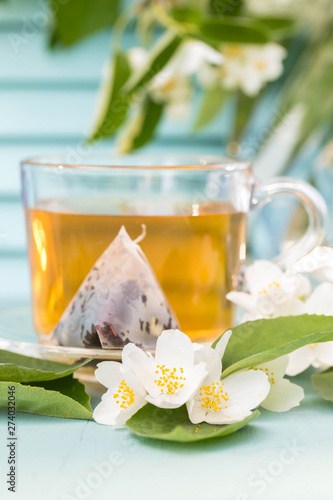 Glass cup with hot jasmine tea on a blue background. The tea bag of tea leaves is taken out from a cup and lies on a saucer. In the foreground jasmine flowers