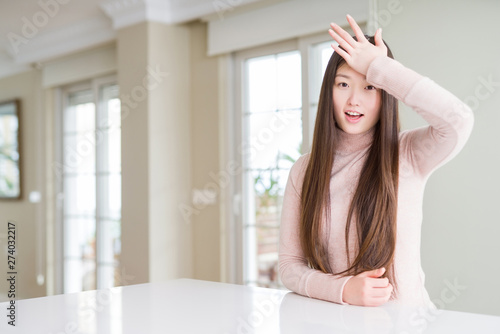 Beautiful Asian woman wearing casual sweater on white table surprised with hand on head for mistake, remember error. Forgot, bad memory concept.