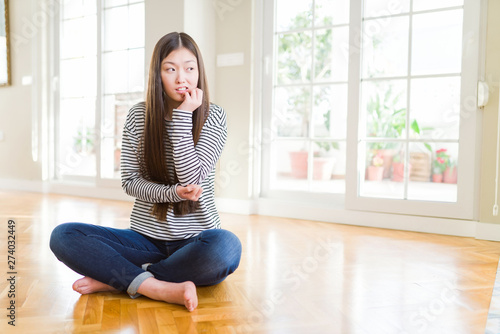 Free Download Beautiful Asian woman sitting barefoot on the floor at home looking stressed and nervous with hands on mouth biting nails. Anxiety problem. - GFXLoad