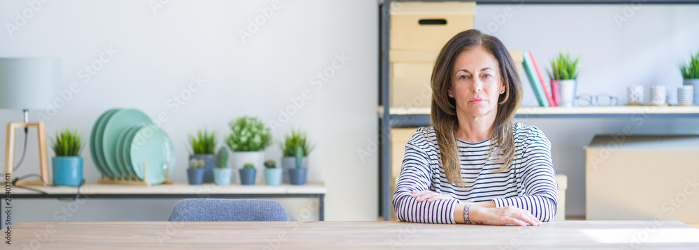Wide angle photo of middle age senior woman sitting at the table at home with serious expression on face. Simple and natural looking at the camera. Wide angle photo of middle age senior woman sitting at the table at home with serious expression on face. Simple and natural looking at the camera.