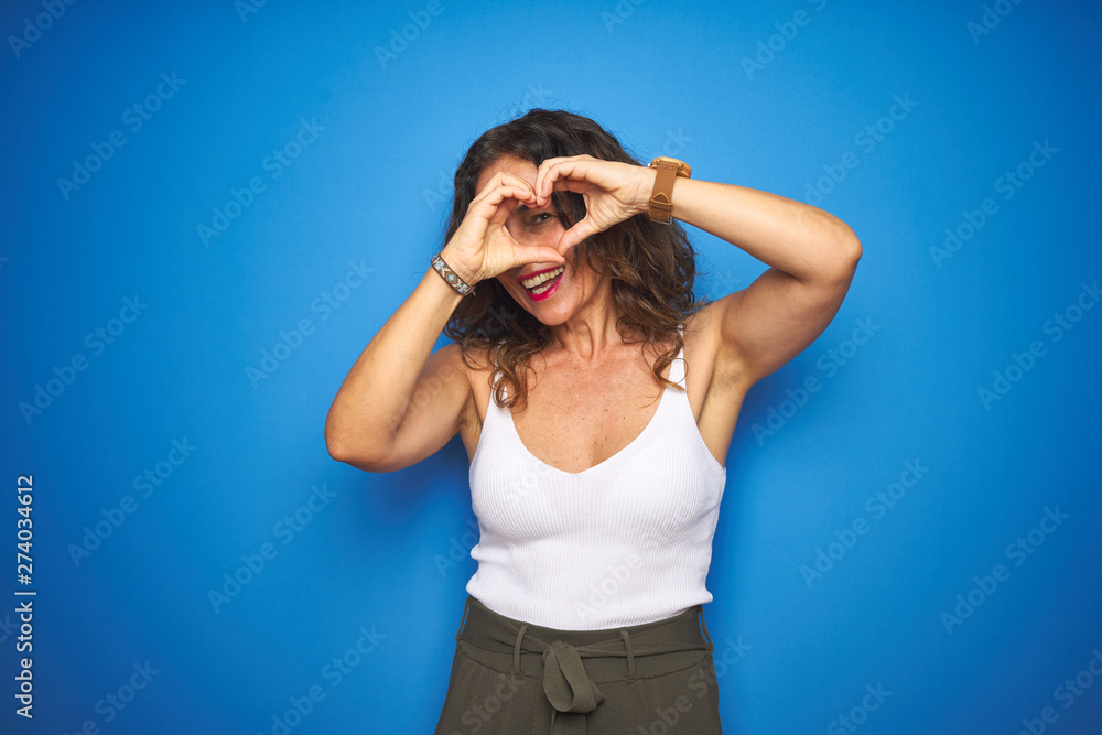 Middle age senior woman with curly hair standing over blue isolated background Doing heart shape with hand and fingers smiling looking through sign Middle age senior woman with curly hair standing over blue isolated background Doing heart shape with hand and fingers smiling looking through sign