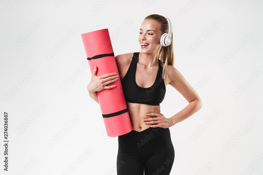 Image of happy caucasian woman wearing tracksuit and headphones carrying fitness mat while doing aerobics Image of happy caucasian woman wearing tracksuit and headphones carrying fitness mat while doing aerobics