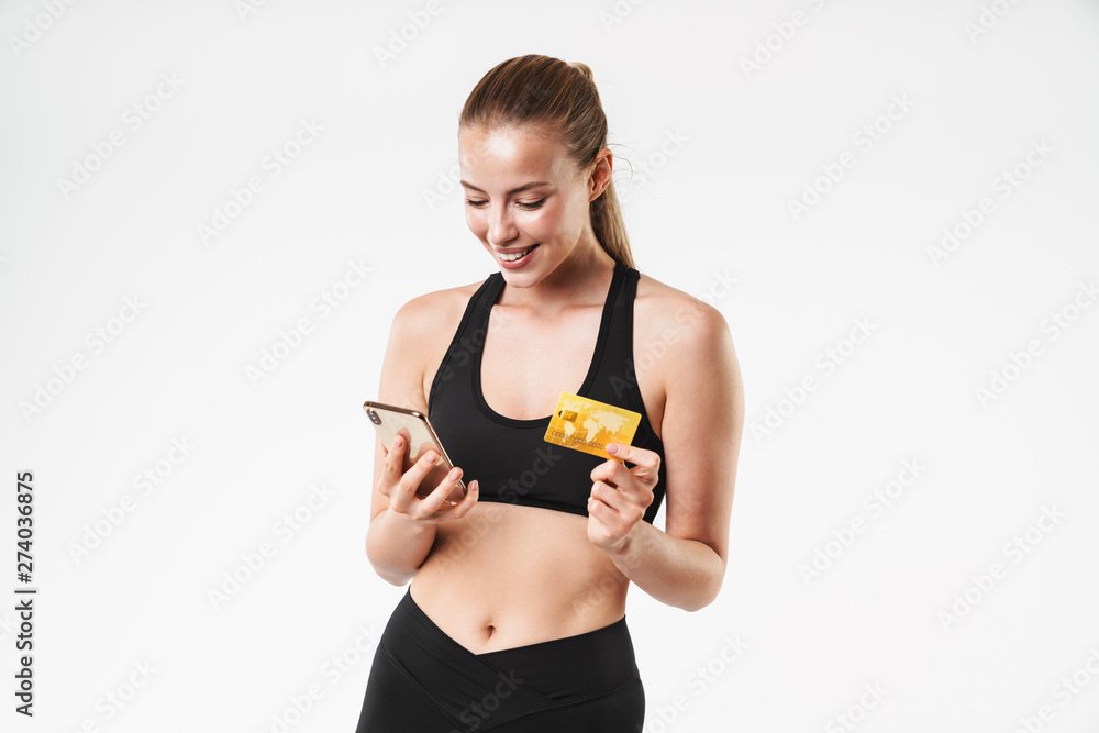 Image of young sporty woman wearing tracksuit rejoicing while holding credit card and cellphone Image of young sporty woman wearing tracksuit rejoicing while holding credit card and cellphone