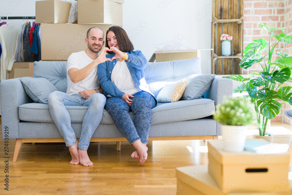 Young couple sitting on the sofa arround cardboard boxes moving to a new house smiling in love doing heart symbol shape with hands. Romantic concept. Young couple sitting on the sofa arround cardboard boxes moving to a new house smiling in love doing heart symbol shape with hands. Romantic concept.