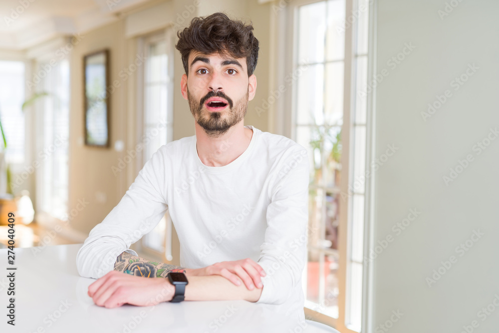 Young man wearing casual shirt sitting on white table afraid and shocked with surprise expression, fear and excited face. Young man wearing casual shirt sitting on white table afraid and shocked with surprise expression, fear and excited face.