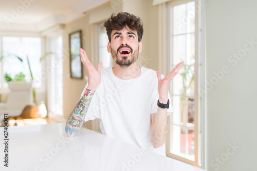 Young man wearing casual t-shirt sitting on white table crazy and mad shouting and yelling with aggressive expression and arms raised. Frustration concept.