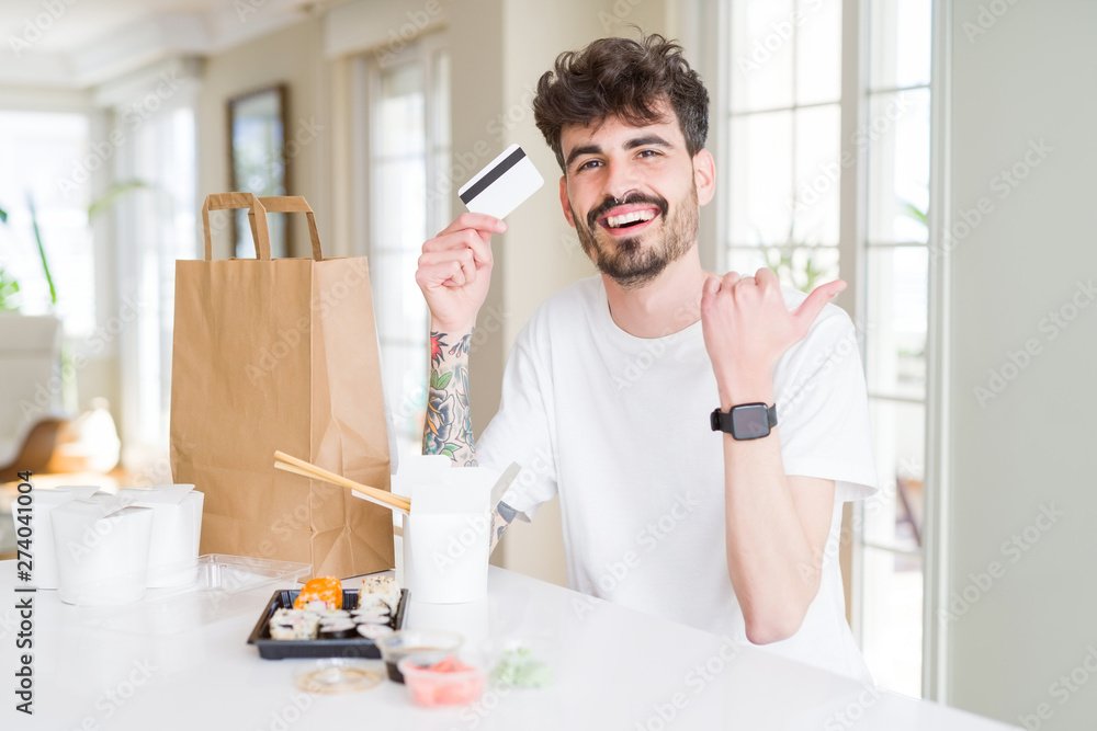 Young man eating asian sushi from home delivery using credit card as payment pointing and showing with thumb up to the side with happy face smiling