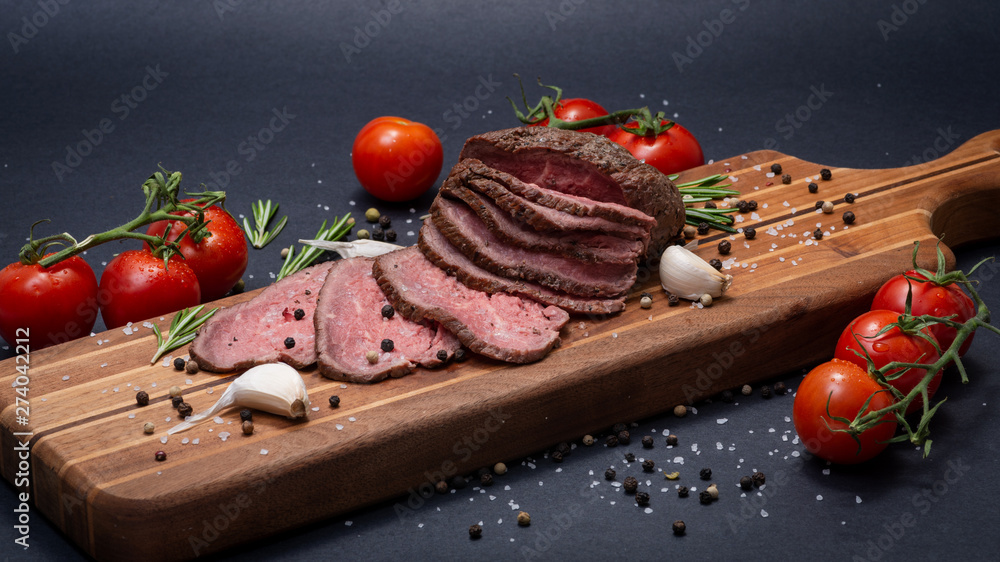 Sliced Grass Fed juicy Corn Roast Beef garnished with Tomatoes, Fresh Rosemary Herb, Garlic and Rainbow Peppercorns on natural wooden cutting board. Sliced Grass Fed juicy Corn Roast Beef garnished with Tomatoes, Fresh Rosemary Herb, Garlic and Rainbow Peppercorns on natural wooden cutting board.