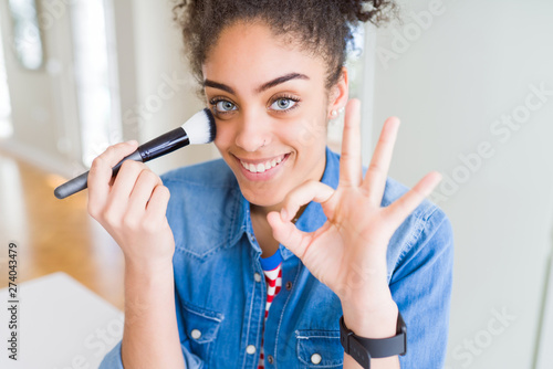 Young african american girl appying blush using make up brush doing ok sign with fingers, excellent symbol