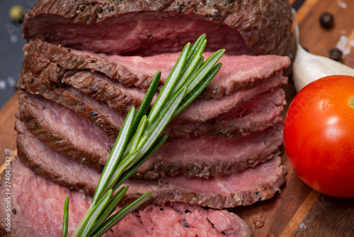 Sliced Grass Fed juicy Corn Roast Beef garnished with Tomatoes, Fresh Rosemary Herb, Garlic and Rainbow Peppercorns on natural wooden cutting board.
