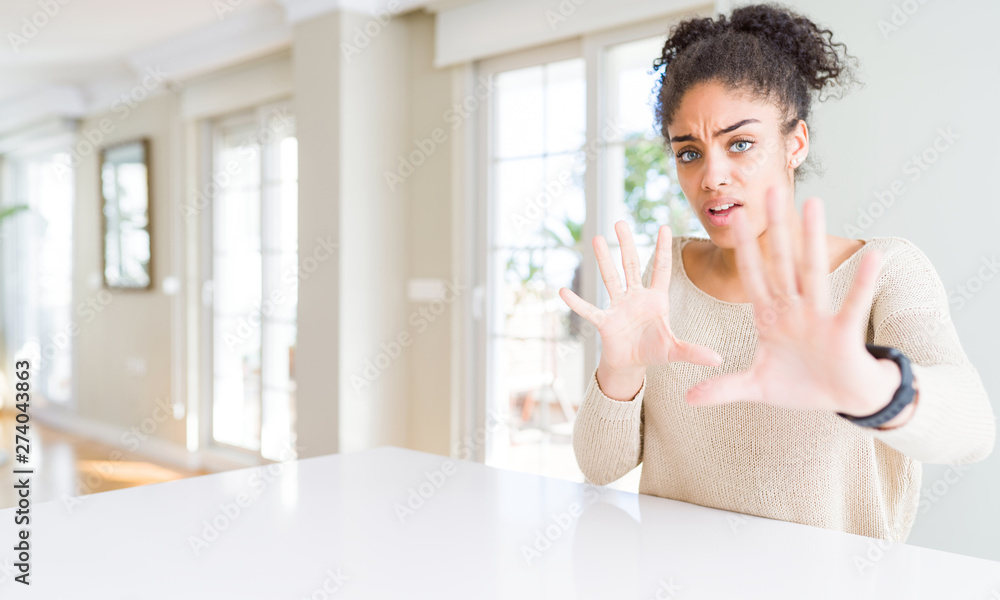 Beautiful young african american woman with afro hair sitting on table at home afraid and terrified with fear expression stop gesture with hands, shouting in shock. Panic concept. Beautiful young african american woman with afro hair sitting on table at home afraid and terrified with fear expression stop gesture with hands, shouting in shock. Panic concept.