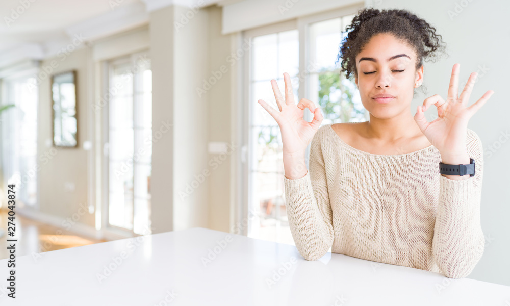 Beautiful young african american woman with afro hair sitting on table at home relax and smiling with eyes closed doing meditation gesture with fingers. Yoga concept. Beautiful young african american woman with afro hair sitting on table at home relax and smiling with eyes closed doing meditation gesture with fingers. Yoga concept.