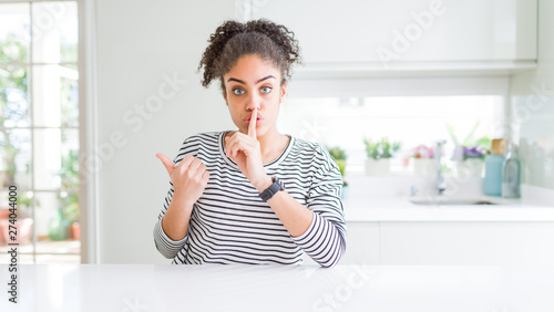 Beautiful african american woman with afro hair wearing casual striped sweater asking to be quiet with finger on lips pointing with hand to the side. Silence and secret concept.