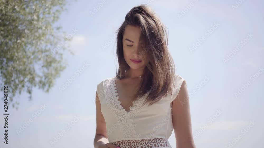 Portrait beautiful young girl with long brunette hair wearing a long white summer fashion dress standing on the waterfront. Leisure pretty woman looking at the camera straightening hair outdoors. Portrait beautiful young girl with long brunette hair wearing a long white summer fashion dress standing on the waterfront. Leisure pretty woman looking at the camera straightening hair outdoors.