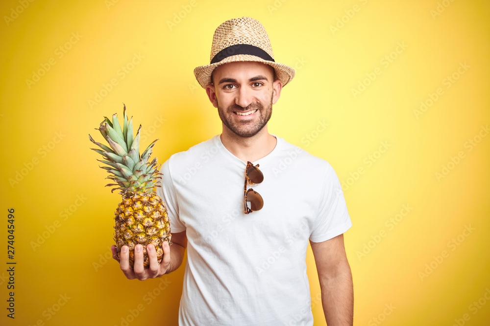 Young man wearing summer hat and holding pineapple over yellow background with a happy face standing and smiling with a confident smile showing teeth Young man wearing summer hat and holding pineapple over yellow background with a happy face standing and smiling with a confident smile showing teeth