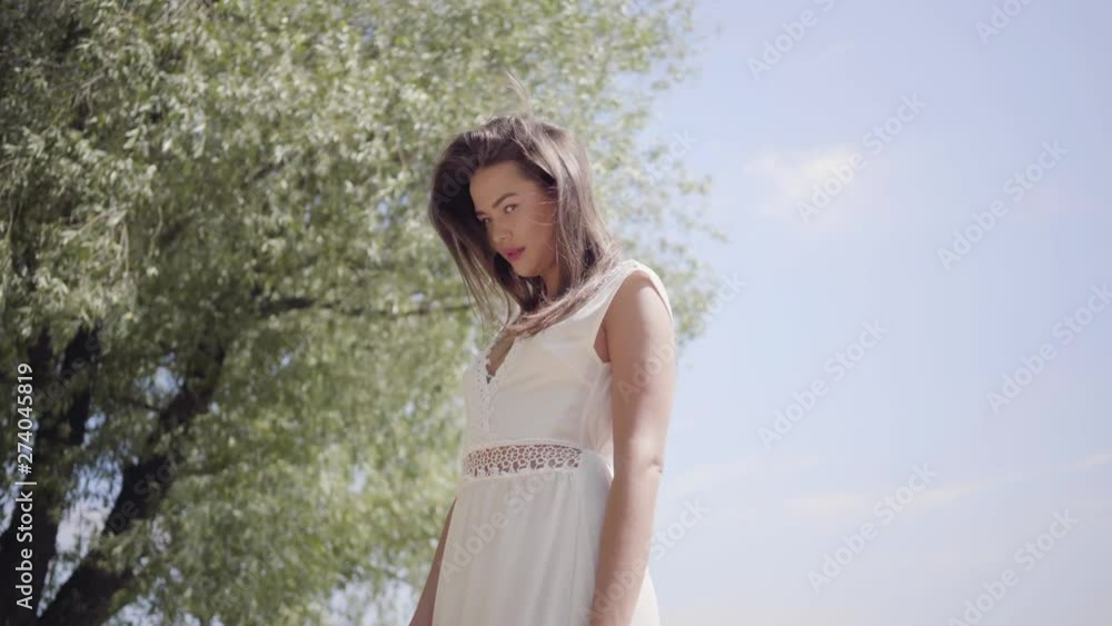 Portrait of lovely young girl with long brunette hair wearing a long white summer fashion dress standing in the park. Leisure pretty woman looking at the camera straightening hair outdoors. Portrait of lovely young girl with long brunette hair wearing a long white summer fashion dress standing in the park. Leisure pretty woman looking at the camera straightening hair outdoors.