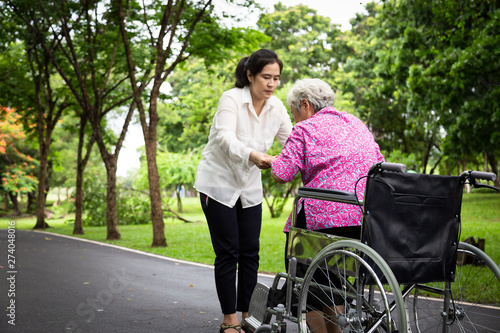 Female caregiver asian or adult nurse support,helping senior woman to stand up from wheelchair in outdoor park,patient mother with her daughter in summer green nature, family ,elderly care concept
