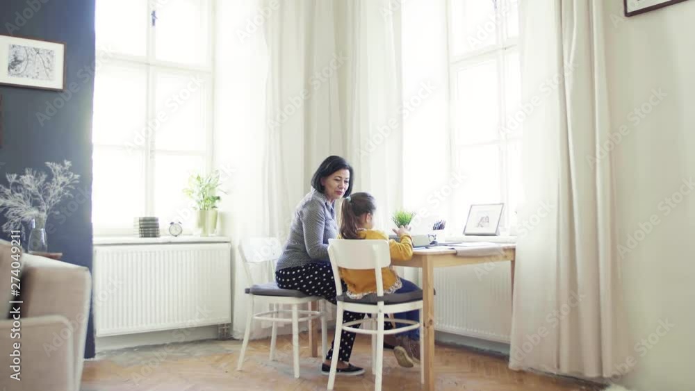 A small girl with grandmother sitting at table at home, using typewriter. A small girl with grandmother sitting at table at home, using typewriter.