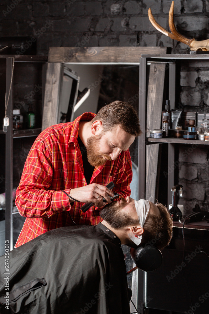 Beard cutting, face care. Barber work with clipper machine in barbershop. Professional trimmer tool cuts beard and hair of young guy in barber shop salon. Beard cutting, face care. Barber work with clipper machine in barbershop. Professional trimmer tool cuts beard and hair of young guy in barber shop salon.