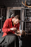 Beard cutting, face care. Barber work with clipper machine in barbershop. Professional trimmer tool cuts beard and hair of young guy in barber shop salon.