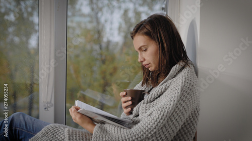 student girl is reading book and sipping hot tea, sitting on a windowsill and wrapped in warm blanket in cold autumn day