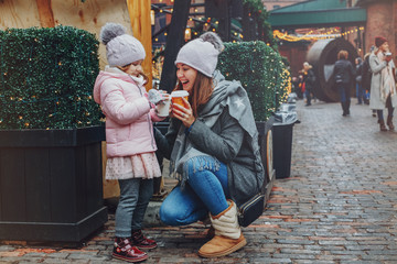 Mother with child girl drinking hot chocolate coffee at Christmas market celebrating New Year holiday. Family outdoor winter activity. Mom and daughter spend time together. Authentic lifestyle.