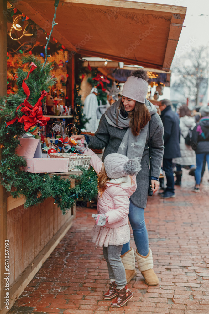 Mother with child girl choosing buying gifts at Christmas market and celebrating New Year holiday. Family outdoor winter activity. Mom and daughter spend time together. Authentic lifestyle.