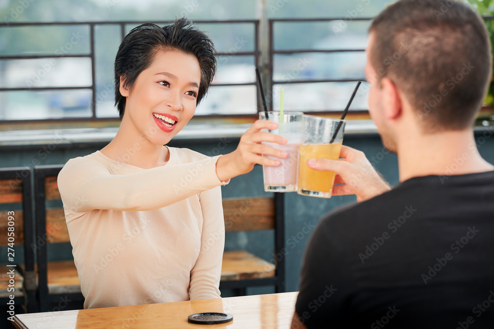 Asian young woman holding glass with nonalcoholic cocktail smiling and toasting with her boyfriend during their dating in outdoor coffee shop Asian young woman holding glass with nonalcoholic cocktail smiling and toasting with her boyfriend during their dating in outdoor coffee shop