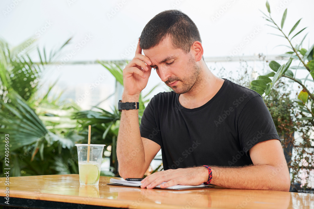 Young businessman sitting at the table with notepad and glass of juice with pensive expression in outdoor coffee shop Young businessman sitting at the table with notepad and glass of juice with pensive expression in outdoor coffee shop