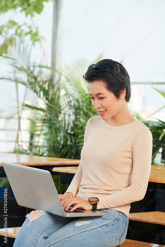 Asian young woman in casual clothing sitting with laptop computer on her knees and communicating online in coffee shop Asian young woman in casual clothing sitting with laptop computer on her knees and communicating online in coffee shop