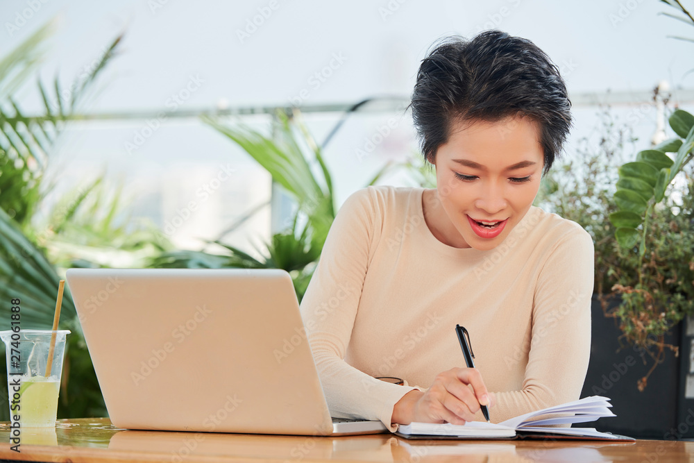 Asian young woman working as a freelancer she sitting at the table with laptop and making notes in her notepad in outdoor coffee shop Asian young woman working as a freelancer she sitting at the table with laptop and making notes in her notepad in outdoor coffee shop