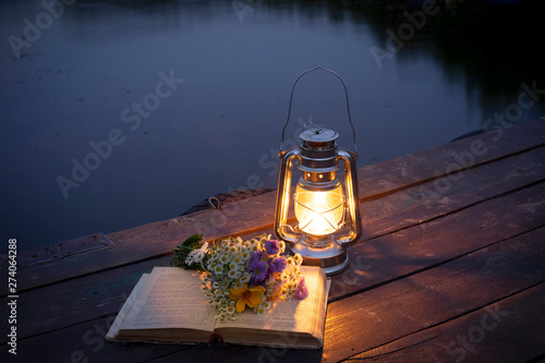 Burning oil lamp, a book, a bouquet of wild flowers on a wooden bridge against the background of a forest lake.